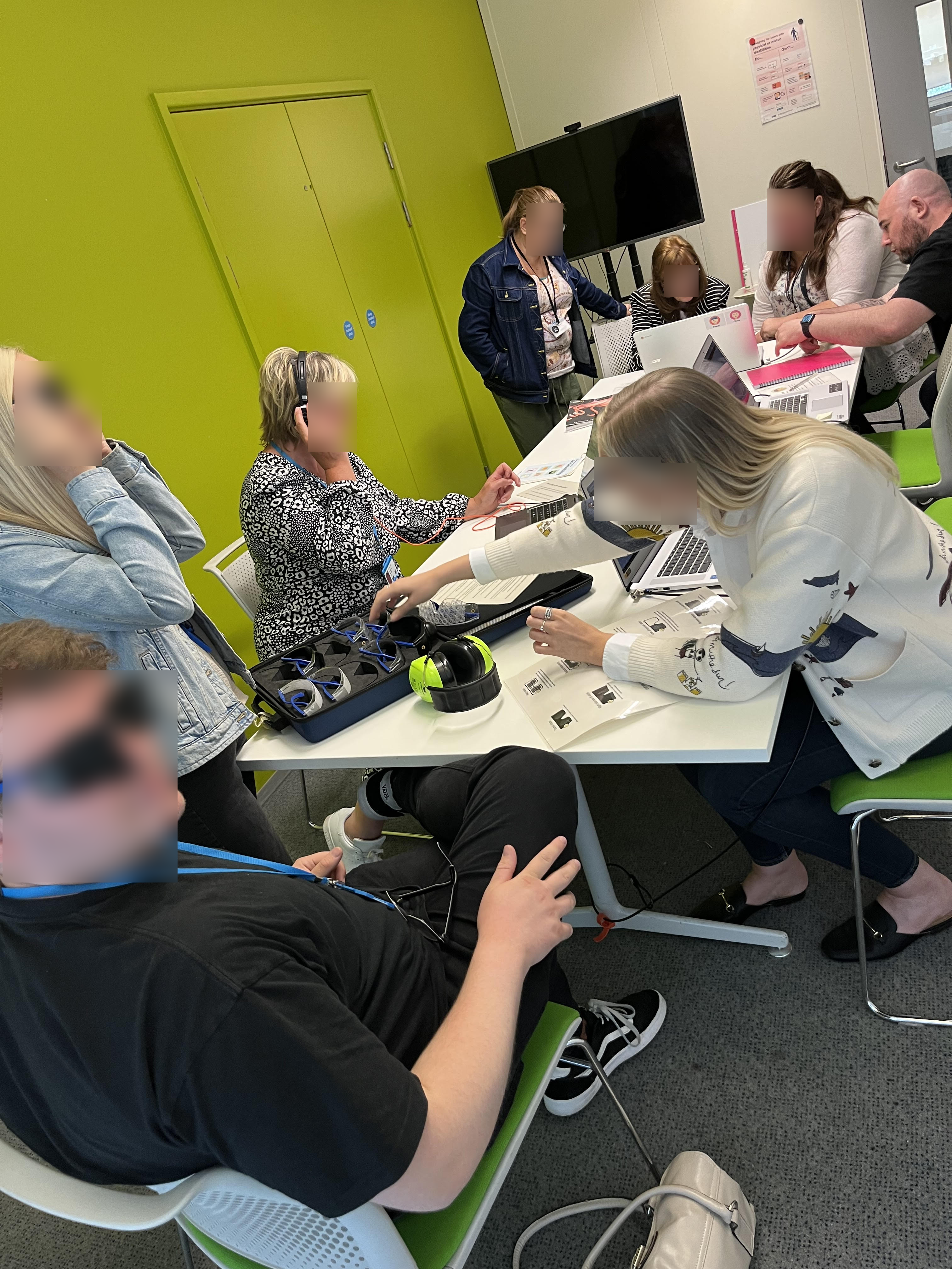 8 people gathered around a table in our empathy hub, some are wearing simulation glasses, one has ear defenders on and the rest are gathered around a laptop talking to me